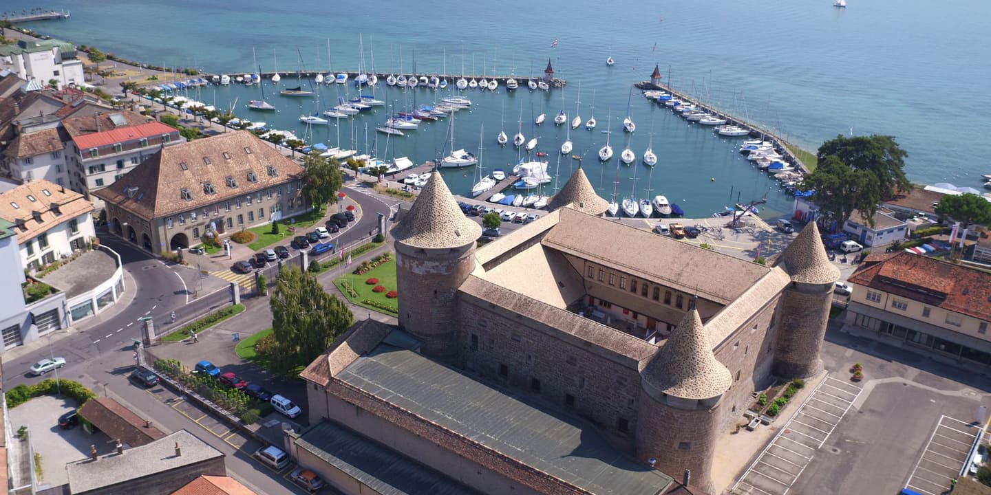 Vue aérienne du château de Morges et de ses quatre tours d’angle, avec le port, ses voiliers et le lac Léman en arrière-plan.